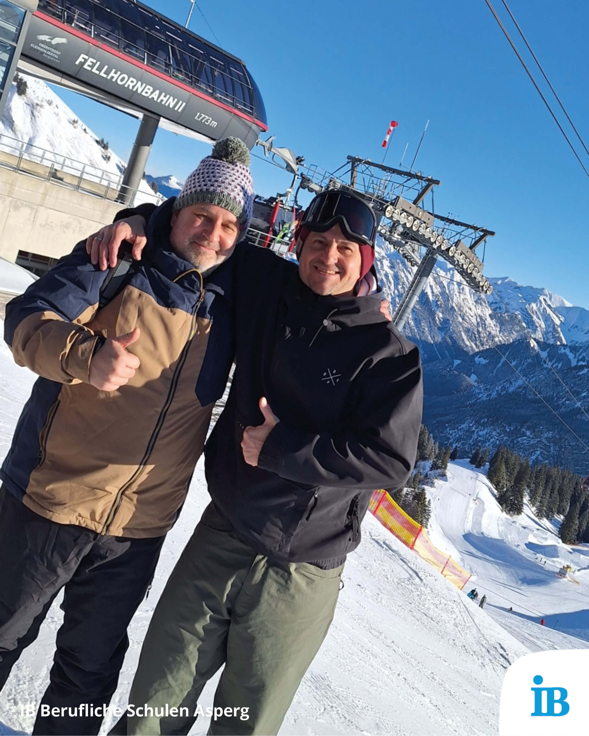 Zwei Männer stehen lächelnd vor einer Seilbahnstation in schneebedeckten Bergen und heben den Daumen hoch.